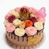 Round chocolate fresh cream cake decorated with chocolate drip, handcrafted fresh cream roses in pink, ivory, burgundy, peach and orange, a golden crown topper, and butterfly decorations.