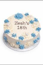 Tall white fresh cream birthday cake decorated with blue and white buttercream flowers, edible pearl accents, and a personalised birthday message.