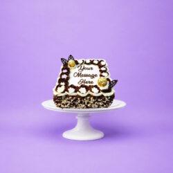 Square fresh cream celebration cake decorated with chocolate and vanilla swirls, white floral piping, Ferrero Rocher chocolates, chocolate leaf decorations, milk and white chocolate shavings, and a personalised message.