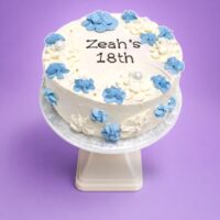 Top view of a tall fresh cream birthday cake featuring blue and white floral decorations, pearl accents, and a personalised birthday message.
