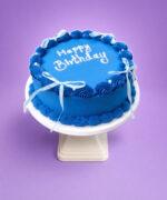 Top view of a royal blue fresh cream birthday cake featuring shell borders, white ribbon bows, pearl accents, and custom birthday writing.