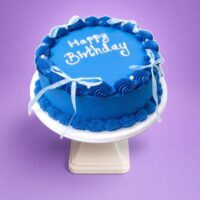 Top view of a royal blue fresh cream birthday cake featuring shell borders, white ribbon bows, pearl accents, and custom birthday writing.