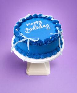 Top view of a royal blue fresh cream birthday cake featuring shell borders, white ribbon bows, pearl accents, and custom birthday writing.