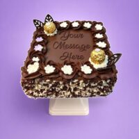 Square chocolate celebration cake with smooth chocolate frosting, Ferrero Rocher chocolates, chocolate leaf decorations, white cream flowers, mixed chocolate curls around the sides, and a personalised message area in the centre.