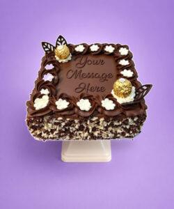 Square chocolate celebration cake with smooth chocolate frosting, Ferrero Rocher chocolates, chocolate leaf decorations, white cream flowers, mixed chocolate curls around the sides, and a personalised message area in the centre.