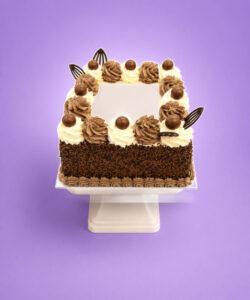 Square chocolate celebration cake with smooth vanilla cream frosting, chocolate buttercream swirls, chocolate vermicelli-coated sides, chocolate decorations, chocolate malt balls, chocolate shell border, and a large blank centre ready for a personalised edible photo or custom message.
