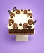 Top view of a square chocolate celebration cake featuring alternating vanilla and chocolate buttercream swirls, chocolate malt balls, decorative chocolate accents, and a smooth blank centre designed for a personalised edible photo or celebration message.