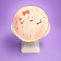 Round pink tooth-themed birthday cake with adorable tooth character and personalized message