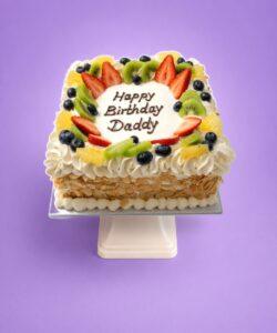 Egg-free fresh fruit celebration cake topped with strawberries, kiwi, blueberries, orange segments, cream piping and almond flakes with a personalised birthday message.