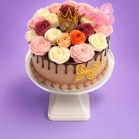 Round chocolate fresh cream cake decorated with chocolate drip, handcrafted fresh cream roses in pink, ivory, burgundy, peach and orange, a golden crown topper, and butterfly decorations.