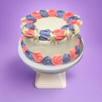 Round fresh cream cake decorated with pink and lavender fresh cream rosettes, white floral accents, textured white sides, and space for a personalised message.
