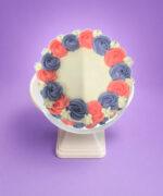 Top view of a white fresh cream celebration cake featuring pink and lavender fresh cream rosettes arranged around the edge with white floral accents and a personalised message area.