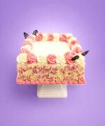 Pink and white celebration cake decorated with whipped frosting swirls, pink and white chocolate curls around the sides, chocolate leaf decorations, and a smooth personalised message area for birthdays and special occasions.