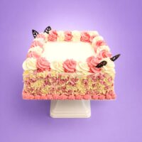 Pink and white celebration cake decorated with whipped frosting swirls, pink and white chocolate curls around the sides, chocolate leaf decorations, and a smooth personalised message area for birthdays and special occasions.