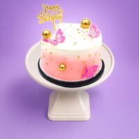 Round Egg Free pink ombre celebration cake decorated with butterfly toppers, edible gold pearls, gold spheres and a Happy Birthday topper.