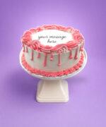 Round white fresh cream birthday cake with pink drip decoration, pink piped borders, textured sides, and a personalised message.