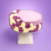 Round fresh cream celebration cake with a pastel lilac finish, purple and ivory rosettes, edible pearl sprinkles, and space for a personalised message.