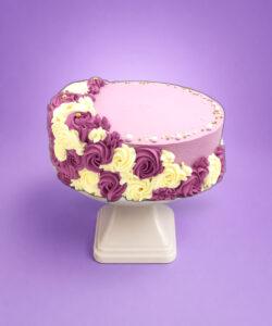 Round fresh cream celebration cake with a pastel lilac finish, purple and ivory rosettes, edible pearl sprinkles, and space for a personalised message.